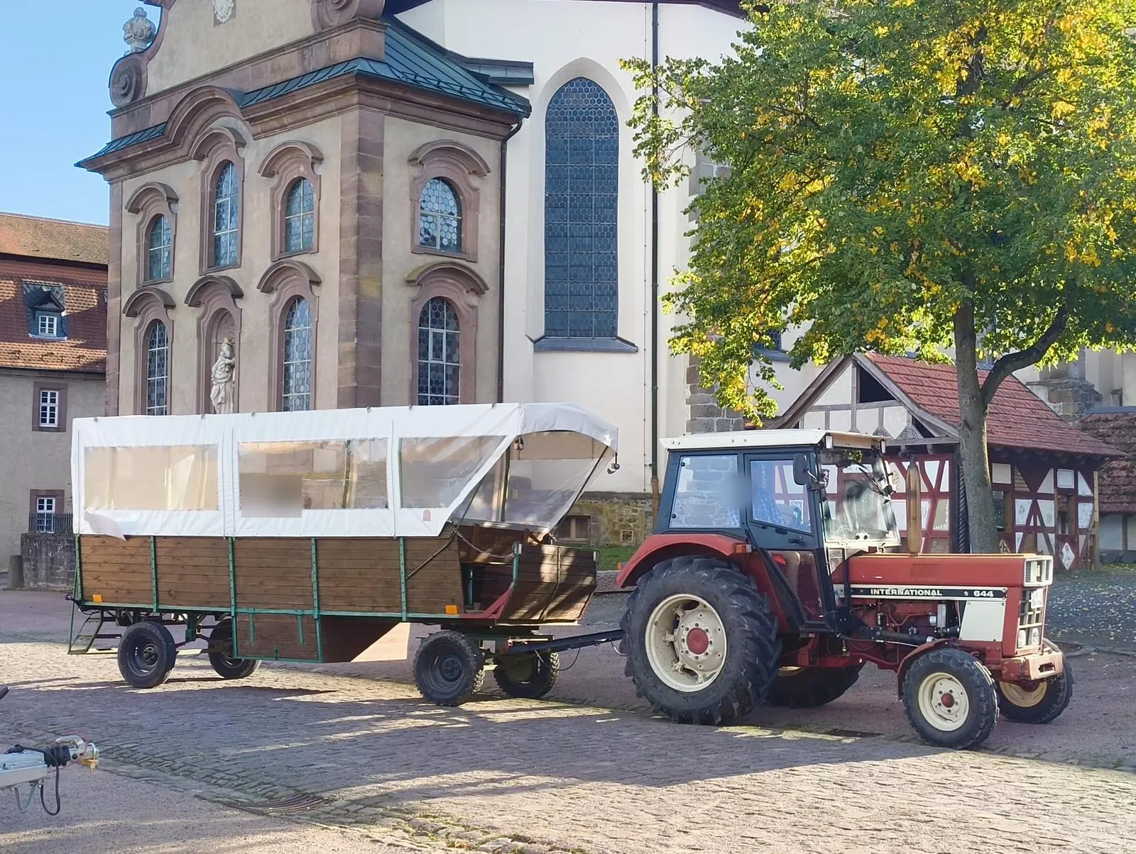 Planwagen mit weißer Plane und Traktor auf einem gepflasterten Platz vor einer Kirche