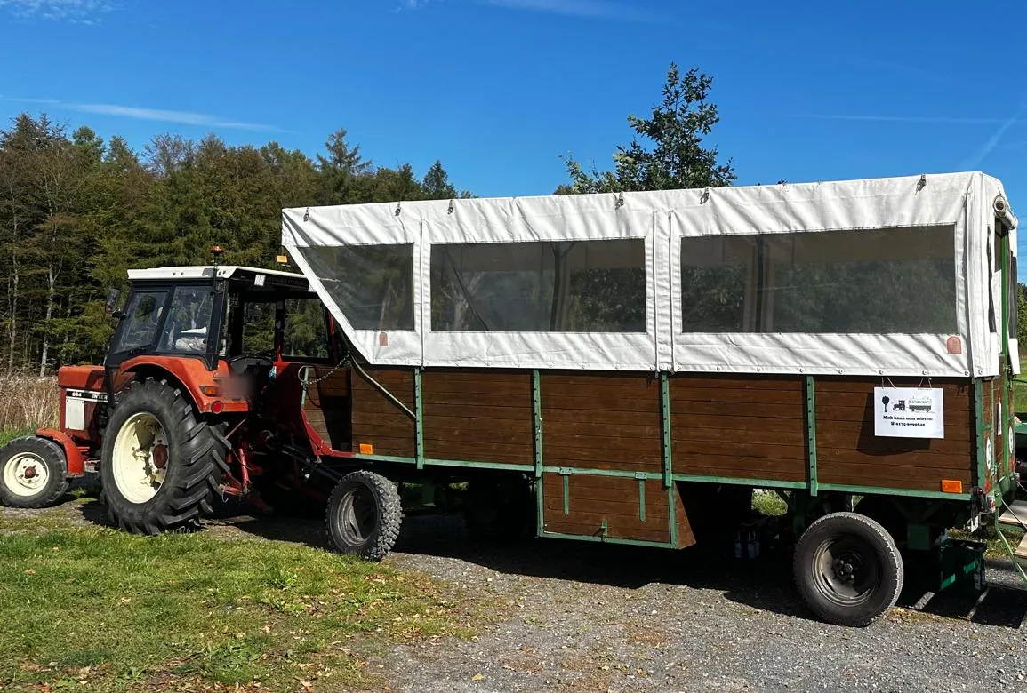 Seitenansicht des Planwagens: Holzaufbau mit weißer Plane und großen Sichtfenstern, davor der Traktor